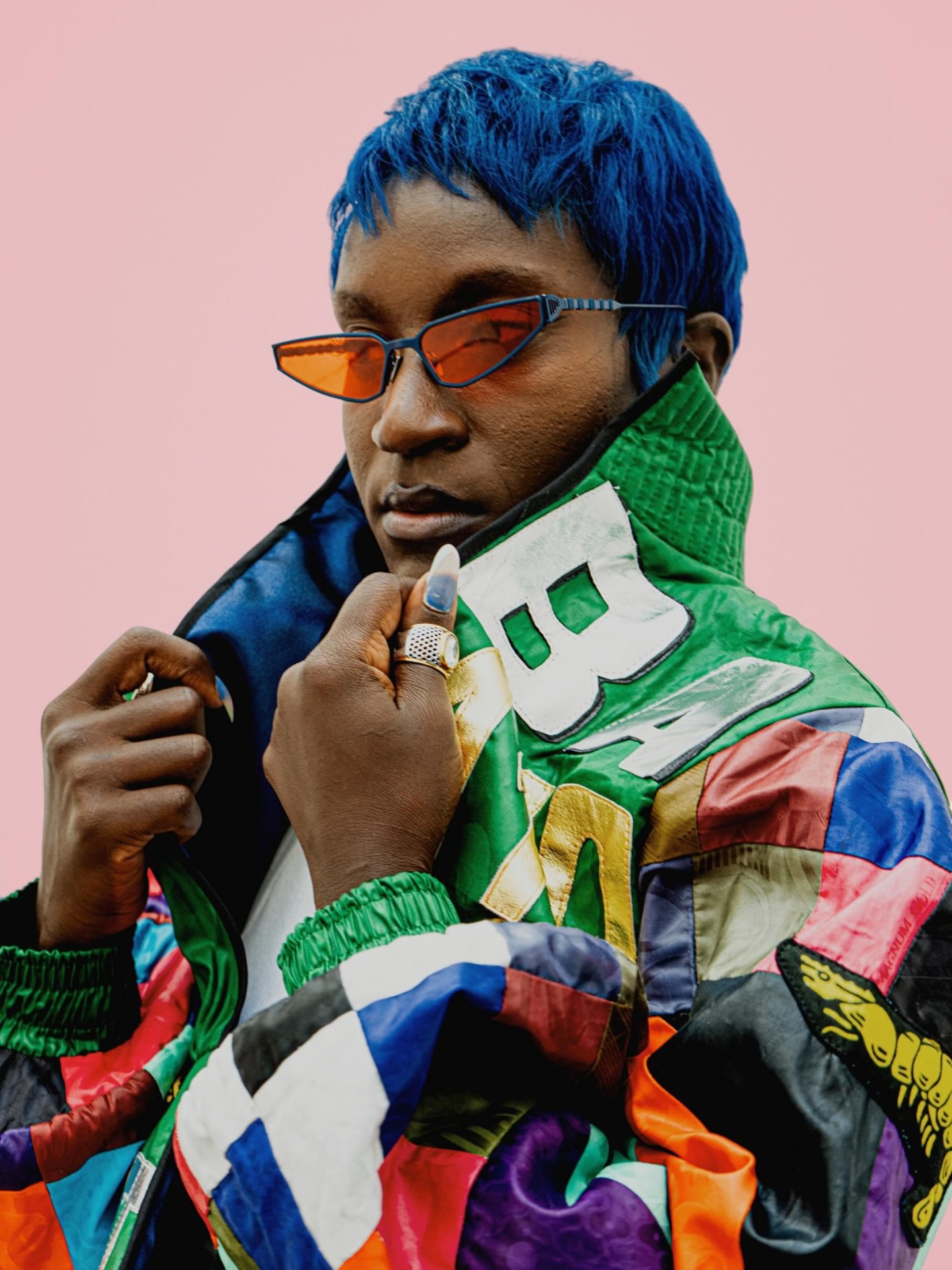 Portrait — cheveux bleus, lunettes orange et blouson patchwork lettering.