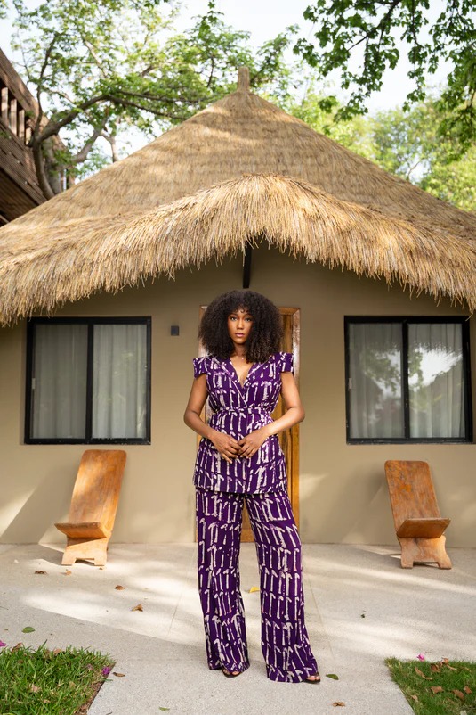 Ensemble pantalon imprimé violet et écru devant une case en paille.